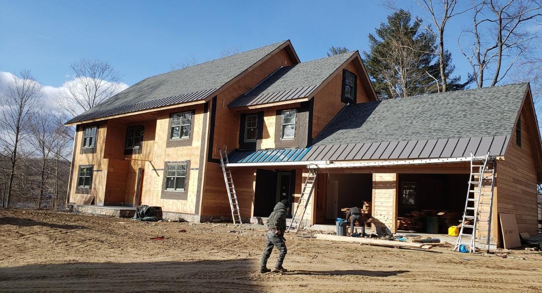Two-story home under construction with dark metal standing seam roofing and gray asphalt shingles on attached garage, wood siding partially installed, ladders leaning against structure, workers on site with building materials scattered across dirt lot