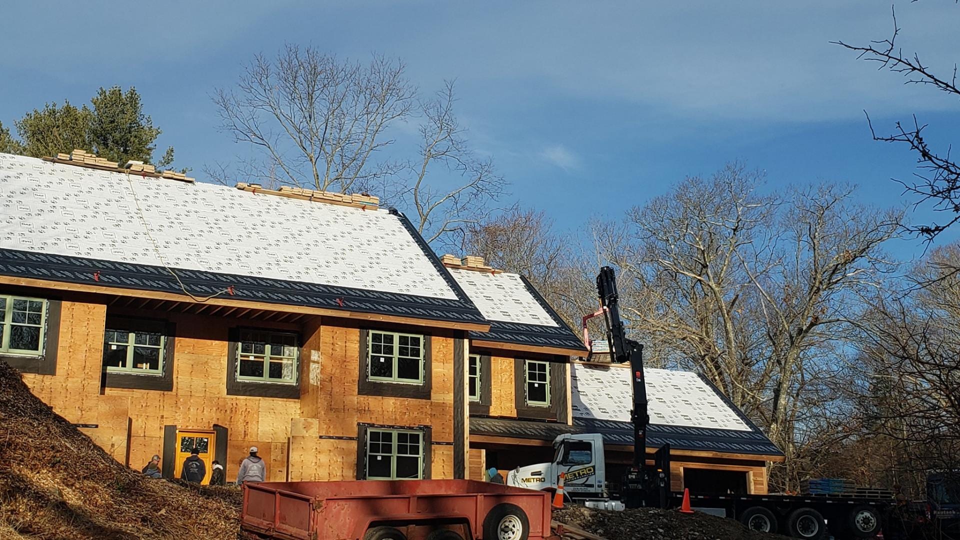 Two-story house under construction with white roof underlayment and stacked shingles, a boom truck crane on site, workers near the entrance, and a dump trailer in the foreground