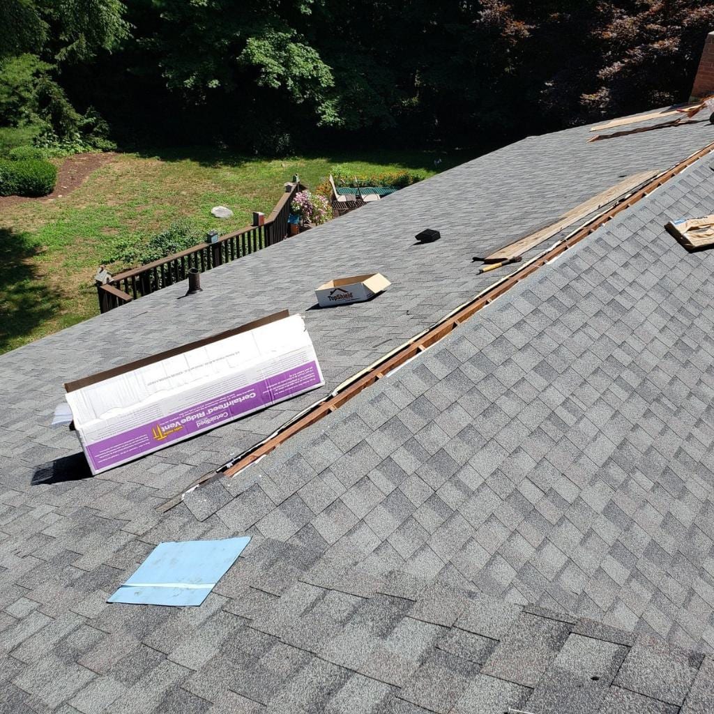 Gray asphalt shingles on a residential roof with an open ridge slot, CertainTeed Ridge Vent packaging, lumber strips, and roofing tools scattered along the peak, with a backyard deck and green trees visible below