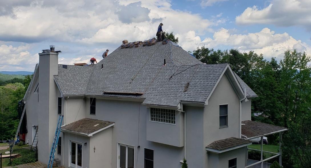 Three roofers removing and replacing shingles on a large hip roof of a two-story gray stucco home, with a blue ladder leaning against the side and green hills visible in the background