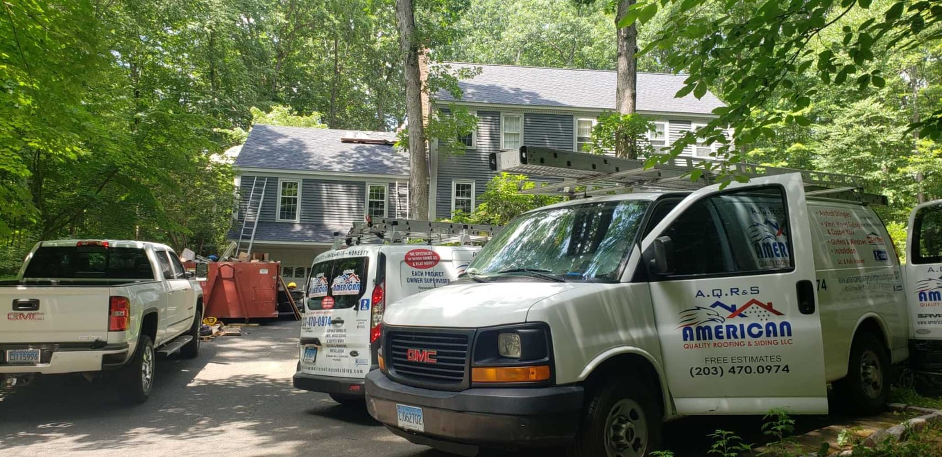 Three white American Quality Roofing and Siding LLC branded GMC vans and a white GMC pickup truck parked in the driveway of a two-story blue-gray colonial home surrounded by mature trees, with a dumpster and ladder visible near the garage during an active roof replacement