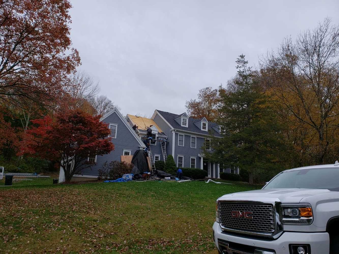 Roofing crew on ladders installing new roof decking on a gray two-story colonial house surrounded by autumn foliage with a white GMC truck in the foreground