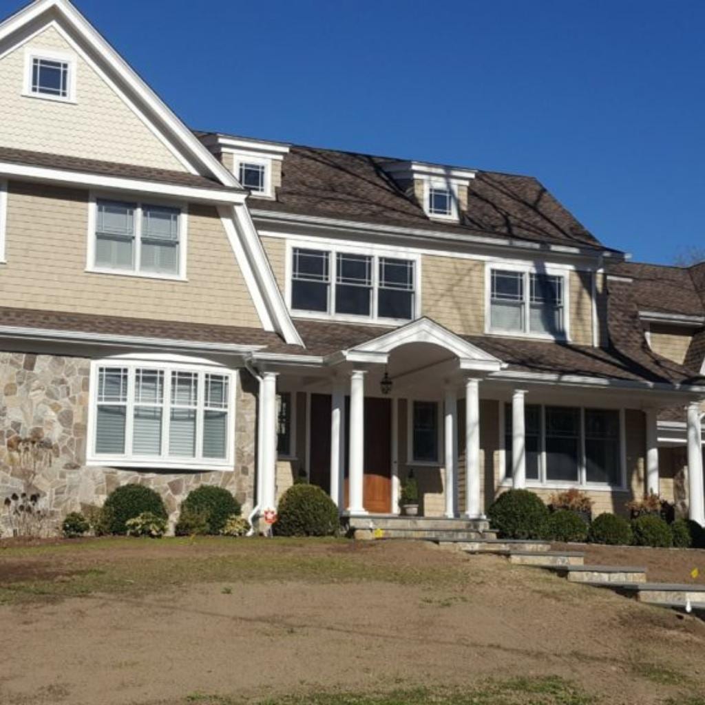 Two-story colonial home with dark brown architectural shingle roof, tan cedar shake siding, stone veneer accent wall, white columned front portico, dormers, and neatly trimmed shrubs along the foundation