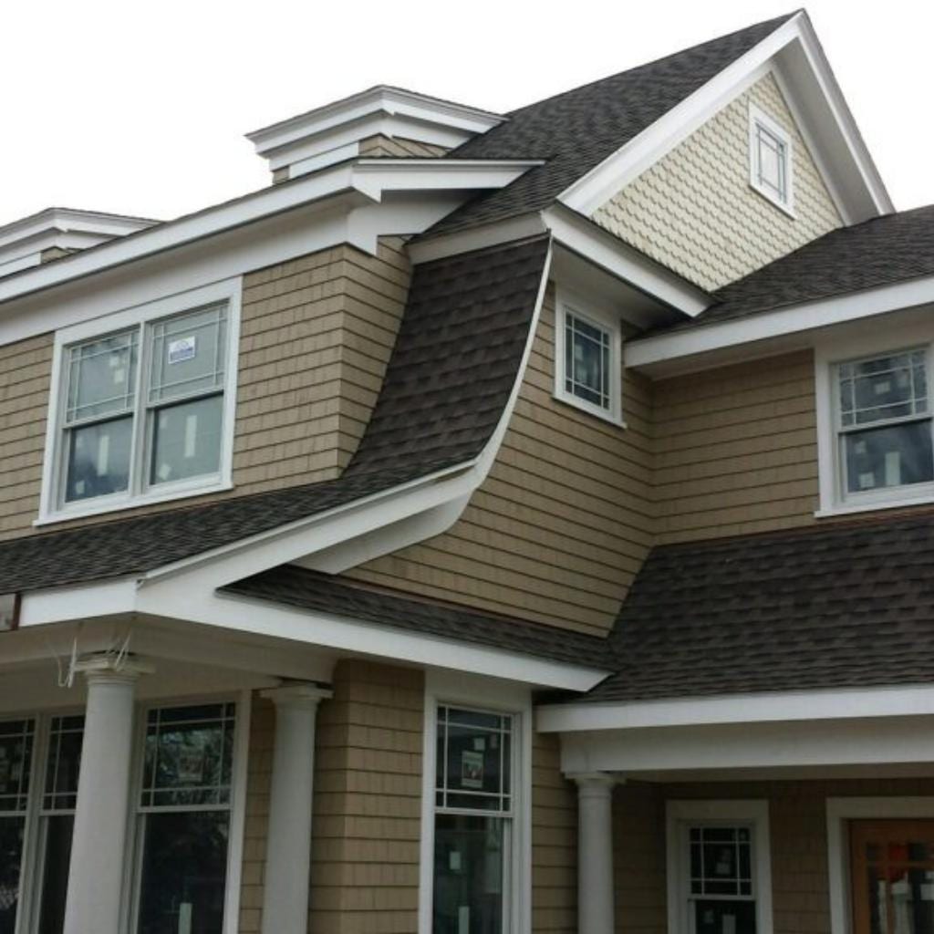 Two-story residential home with dark charcoal architectural shingle roof, tan horizontal vinyl siding, white trim accents, multiple roof dormers, covered front porch with white columns, and large multi-pane windows