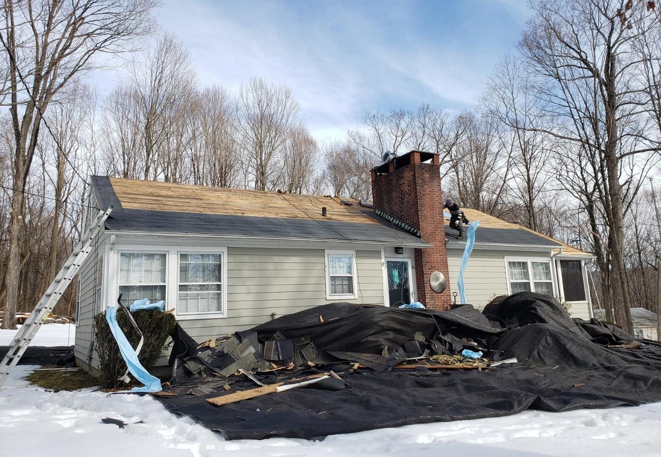 A worker on a ranch-style house roof near a brick chimney removing old shingles, with stripped decking visible, a ladder leaning against the left side, and a large pile of old roofing materials and black tarp on snow-covered ground in front