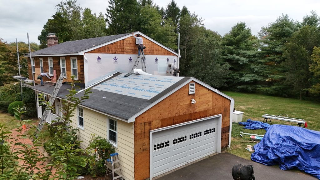 Workers installing house wrap and removing cedar shake siding on a two-story colonial home with attached garage, multiple ladders positioned against the structure, blue tarp covering materials on driveway
