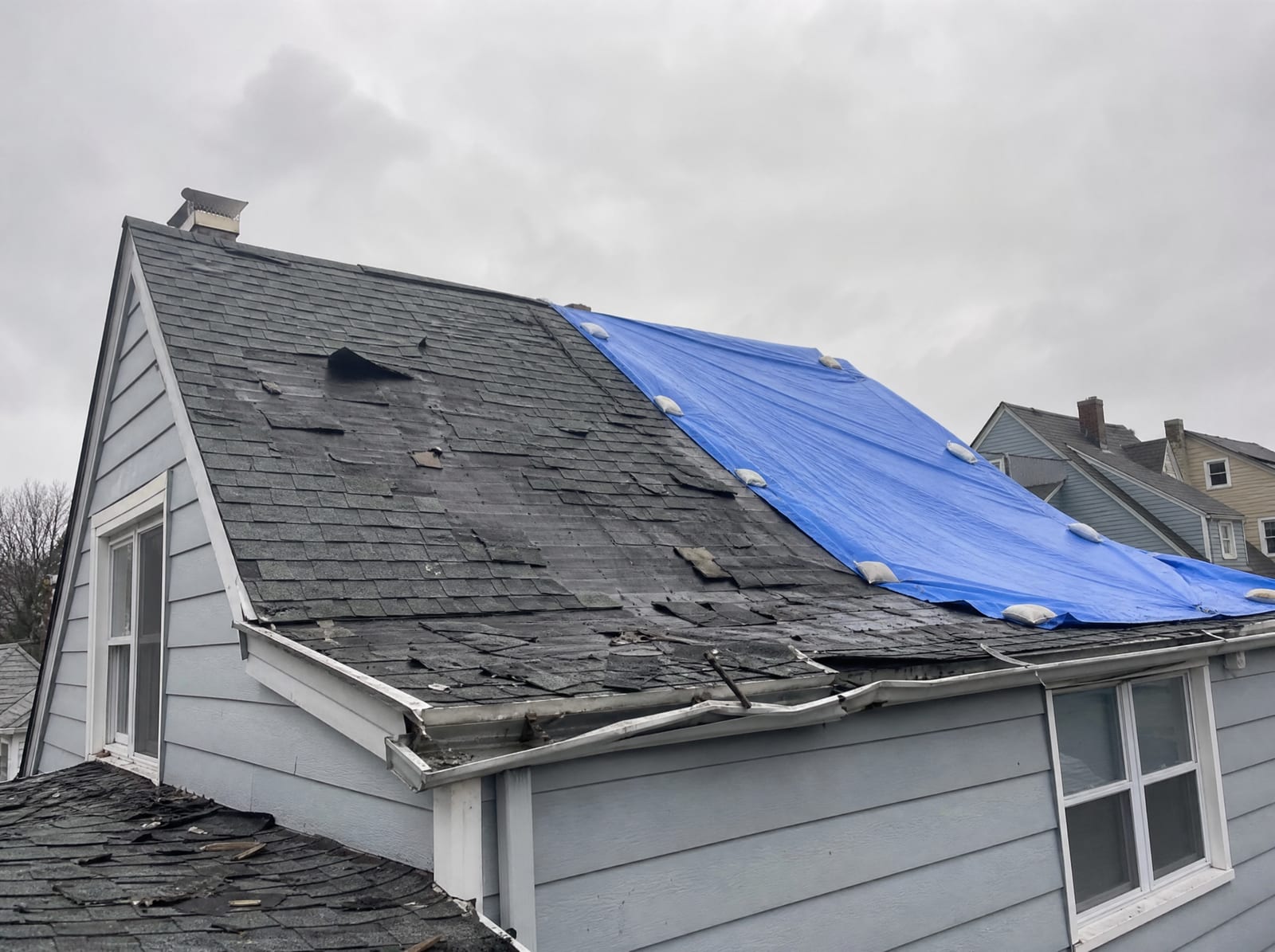 Coastal storm-damaged asphalt shingle roof with wind-torn shingles and a section covered by a secured blue tarp under a gray, windy sky