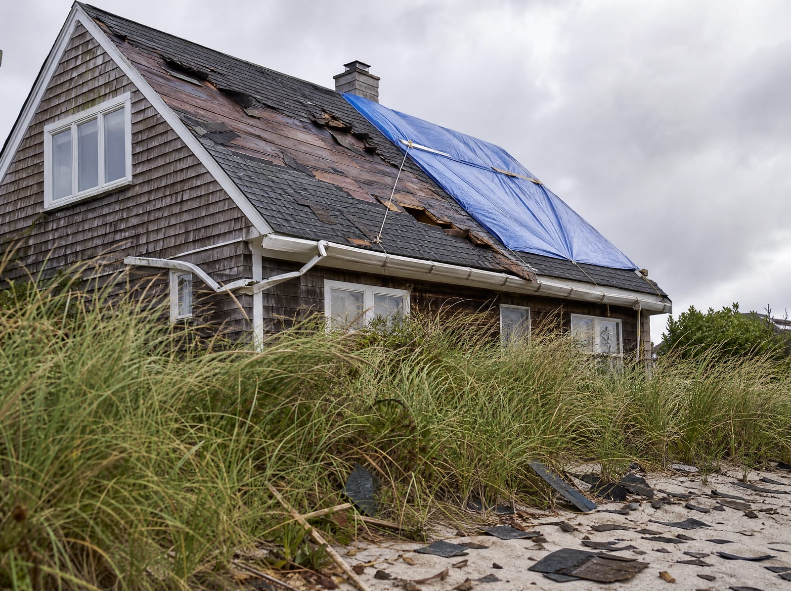 Coastal storm-damaged roof on a Milford Cape home near the shore with wind-torn shingles, a bent gutter, and a secured blue emergency tarp