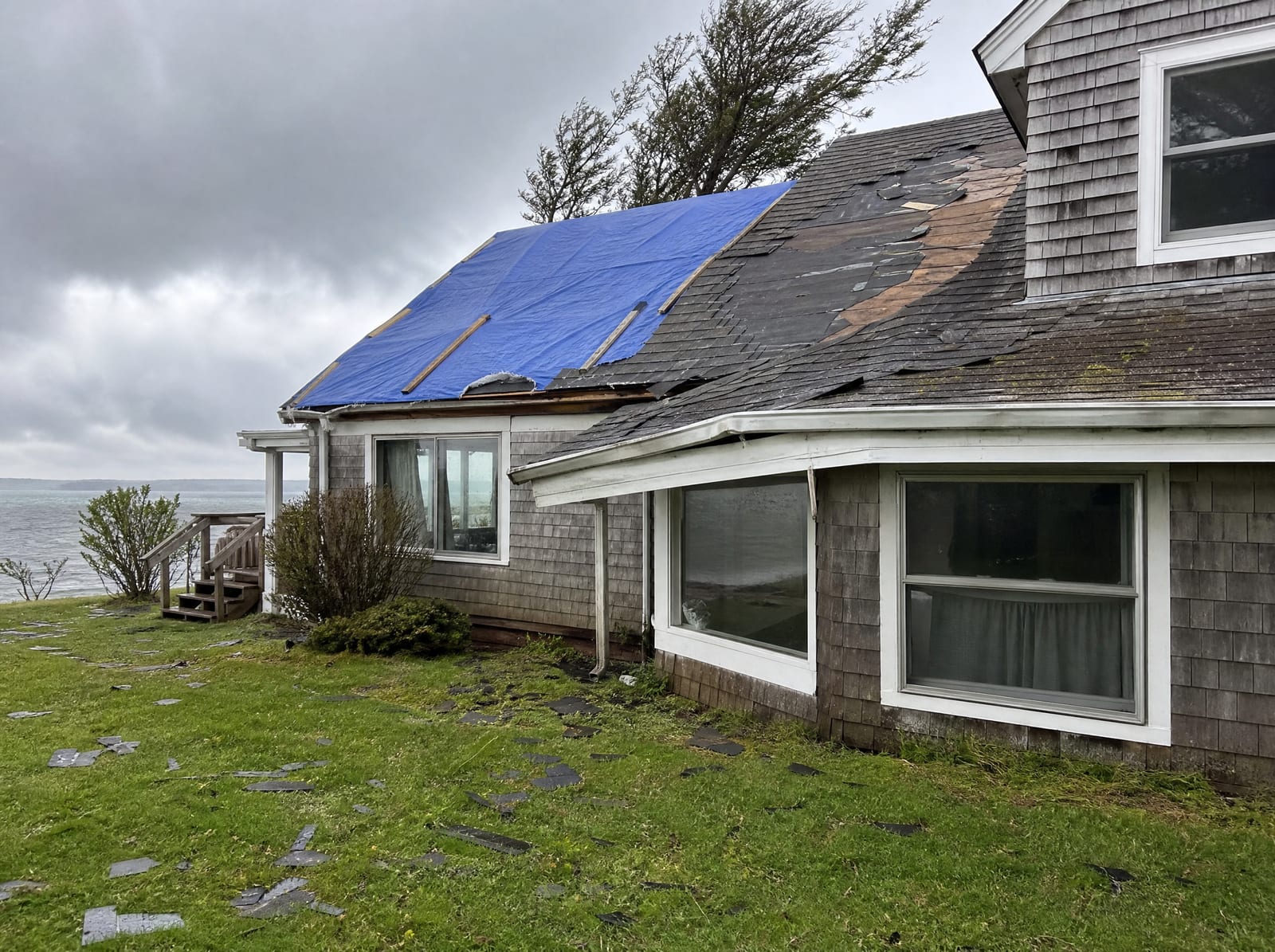 Coastal storm-damaged asphalt shingle roof on a Cape home near the shore with wind-torn shingles, a bent gutter, and a secured blue tarp