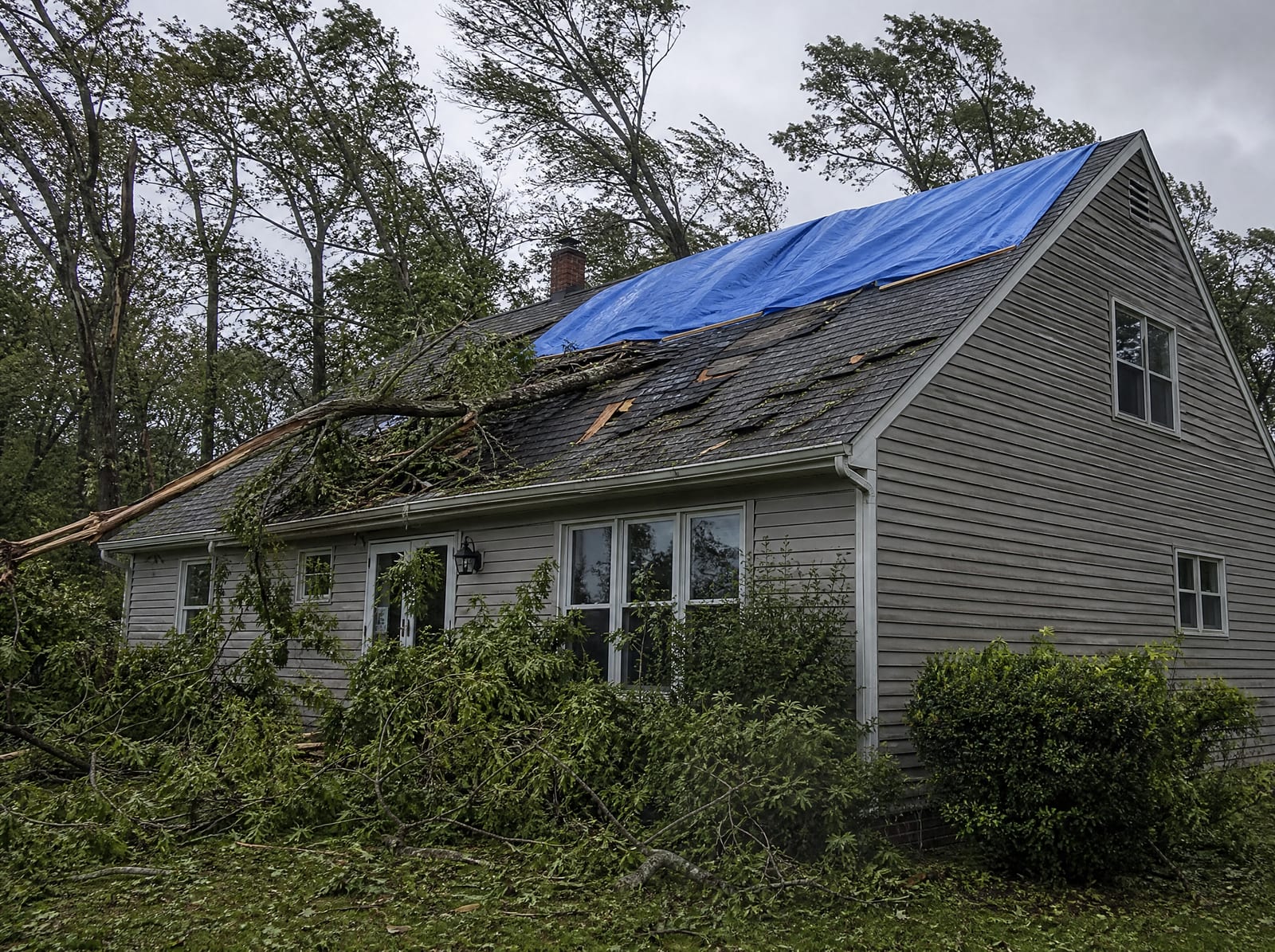 Storm-damaged roof on a wooded Thomaston home with a fallen tree limb, wind-torn shingles, and a secured blue emergency tarp